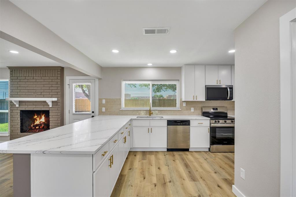 Kitchen featuring white cabinetry, quartz countertops, stainless steel appliances, and wood-finish flooring, complemented by a brick fireplace with a mantel