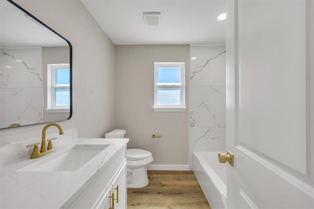 Bathroom featuring a vanity with a stone-surface countertop, gold-tone hardware, a wall-mounted mirror with a black frame, wood-finish flooring, and a tub-shower surround with large-format tile