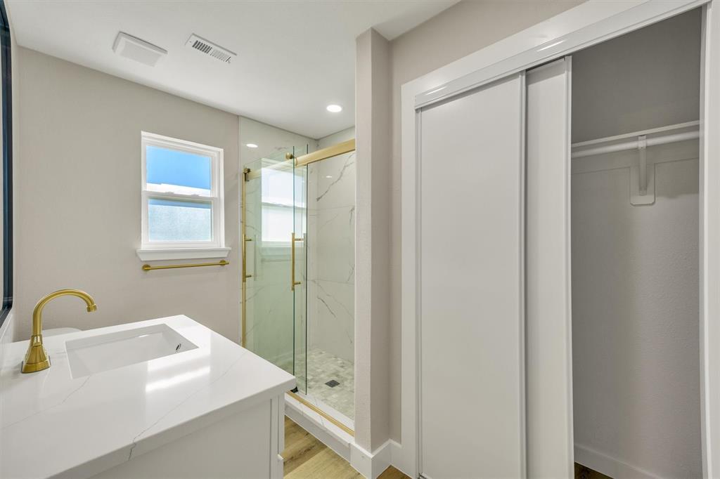 Bathroom featuring a vanity with a white composite countertop, gold-toned faucet, walk-in shower with gold-finished glass enclosure and stone-patterned tile, and a sliding-door closet