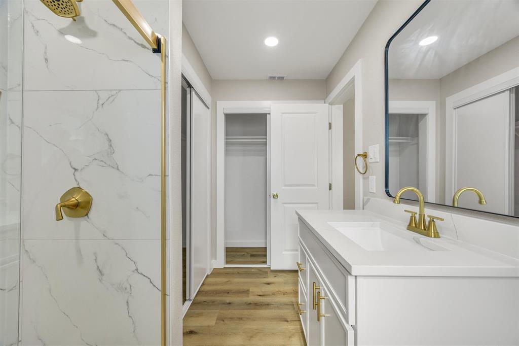 White vanity with solid-surface countertop and gold-toned hardware, gold-toned shower fixtures, large-format stone-patterned wall tile, rectangular mirror with a dark frame, and wood-finish flooring