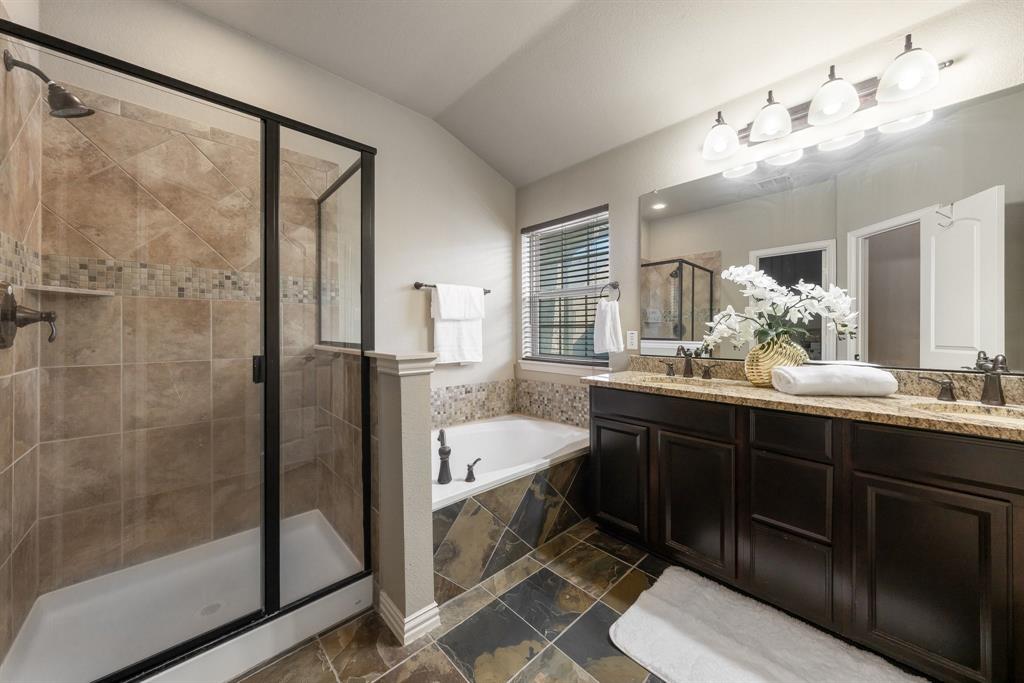 Bathroom featuring a dual-sink vanity with dark cabinetry and stone-finish countertops, a glass-enclosed shower with tiled walls, a deep soaking tub with a tiled surround, and multi-light vanity lighting