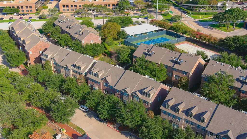 Aerial view of brick-exterior multi-story residences featuring pitched shingle roofing, dormer windows, mature tree-lined landscaping, and a fenced hard-court facility