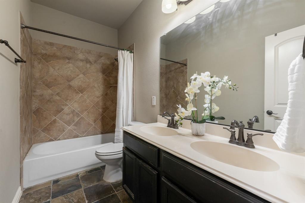 Bathroom featuring a dual-sink vanity with dark cabinetry, stone-finish countertops, tile tub surround set in a diamond pattern, and multi-tone tile flooring