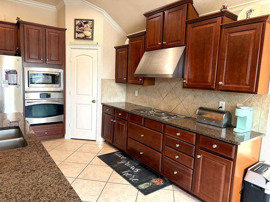 Kitchen featuring rich-toned wood cabinetry, stone countertops, a stainless steel range hood, built-in double oven, and diagonal tile flooring