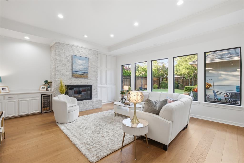 Light pours in through a row of black-framed windows, with a tray ceiling, wide-plank wood floors, and a whitewashed brick fireplace.