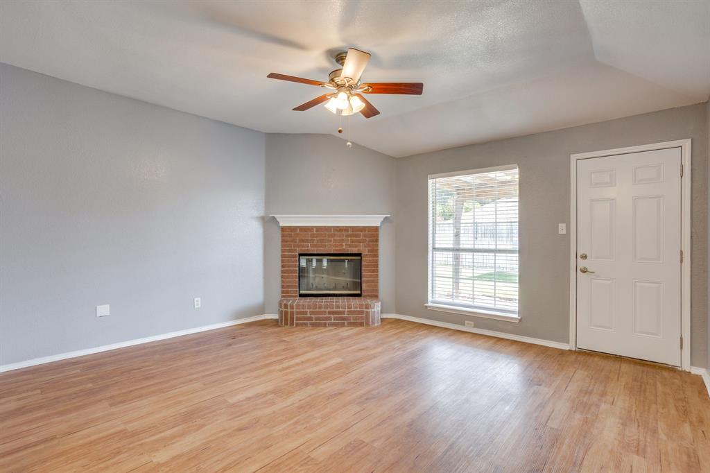 Vaulted ceiling featuring a ceiling fan with integrated lighting, wood-finish flooring, corner brick fireplace with a mantel, and a single-light exterior door