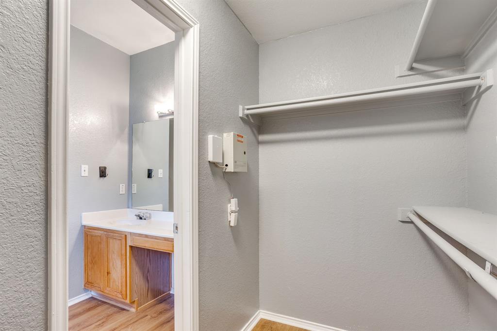 Walk-in closet featuring tiered shelving and wood-finish flooring, complemented by a bathroom area with built-in wood cabinetry, a white countertop, and wall-mounted vanity mirror