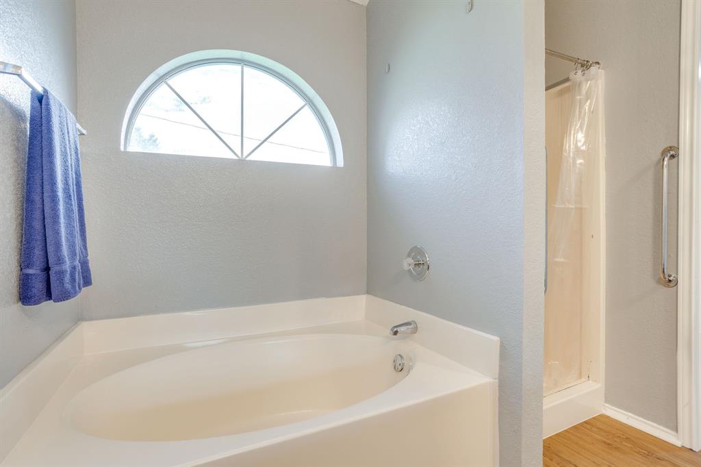 Oval soaking tub with integrated surround, arched window, wood-finish flooring, and shower enclosure with a wall-mounted grab bar
