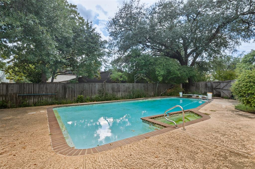 In-ground swimming pool featuring a brick-paver border, a dedicated spa area, metal handrails, and a concrete deck enclosed by a wood privacy fence with mature tree canopy