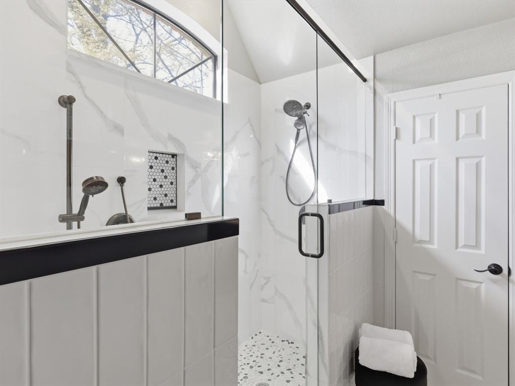 Walk-in shower featuring floor-to-ceiling white tile with veining, glass enclosure with black hardware, built-in mosaic tile niche, dual shower heads, and pebble-style flooring