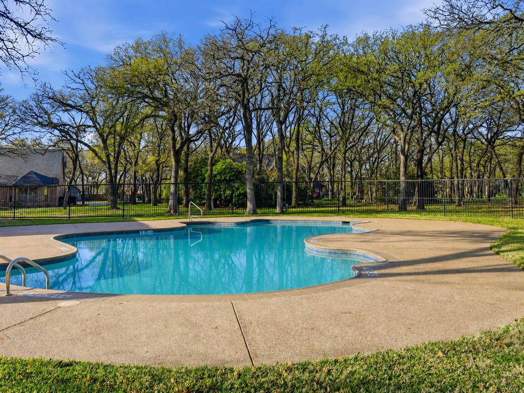 In-ground swimming pool featuring a textured concrete deck, metal perimeter fencing, and mature tree canopy backdrop