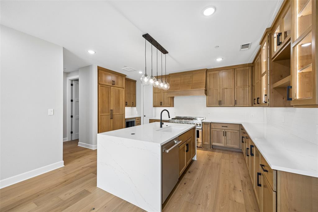 Kitchen featuring custom wood cabinetry, an expansive center island with quartz countertops, integrated professional-grade gas range, wood-finish flooring, and linear multi-pendant lighting