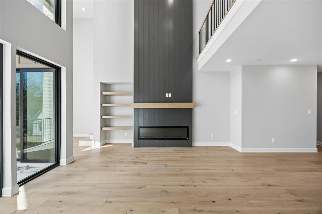 Double-height ceilings, linear fireplace with floor-to-ceiling vertical plank surround, wood-finish flooring, built-in recessed shelving, and mezzanine railing
