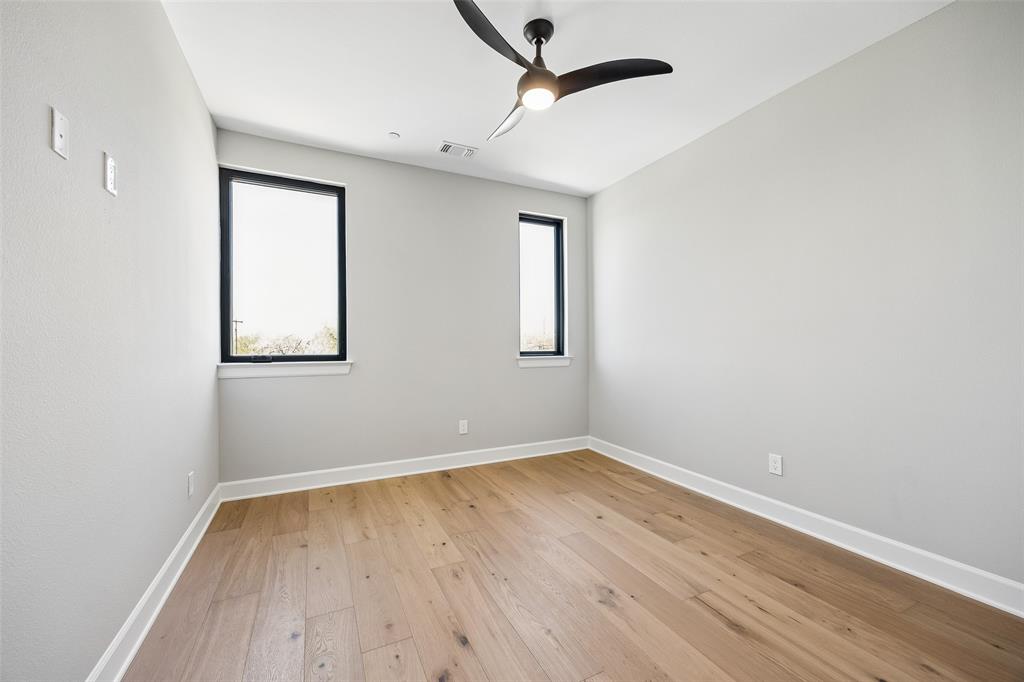 Neutral interior featuring wood-finish flooring, two dual-pane windows with black frames, and a modern three-blade ceiling fan with integrated light