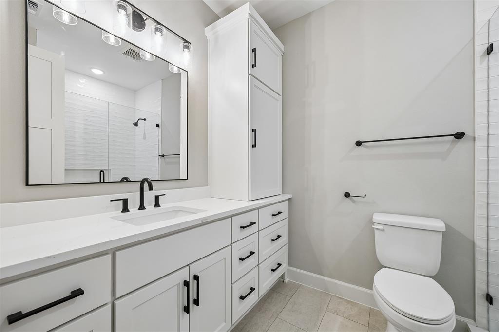 White cabinetry with matte black hardware, stone-surface countertop, rectangular undermount sink, built-in storage tower, and a framed mirror with integrated lighting