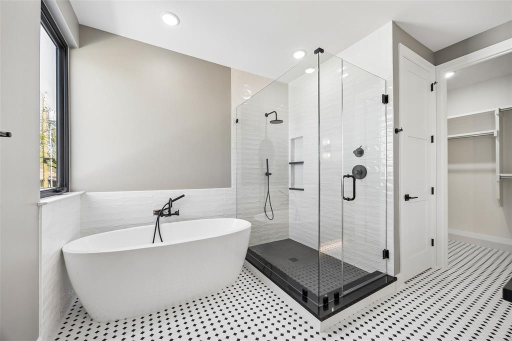 Freestanding soaking tub with floor-mounted faucet, glass-enclosed shower with dark tiled base and recessed shelving, and patterned mosaic tile flooring