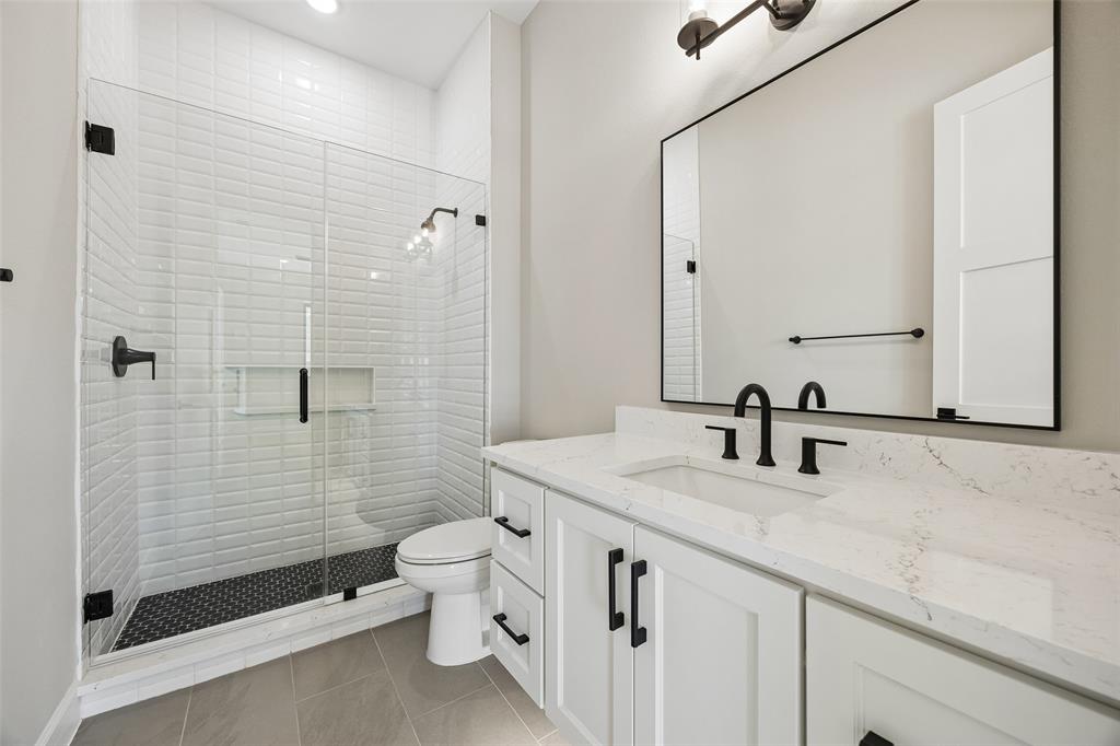 Bathroom featuring a walk-in shower with white subway tile walls and dark patterned flooring, light-toned cabinetry with a solid surface countertop, an undermount sink, and matte black fixtures