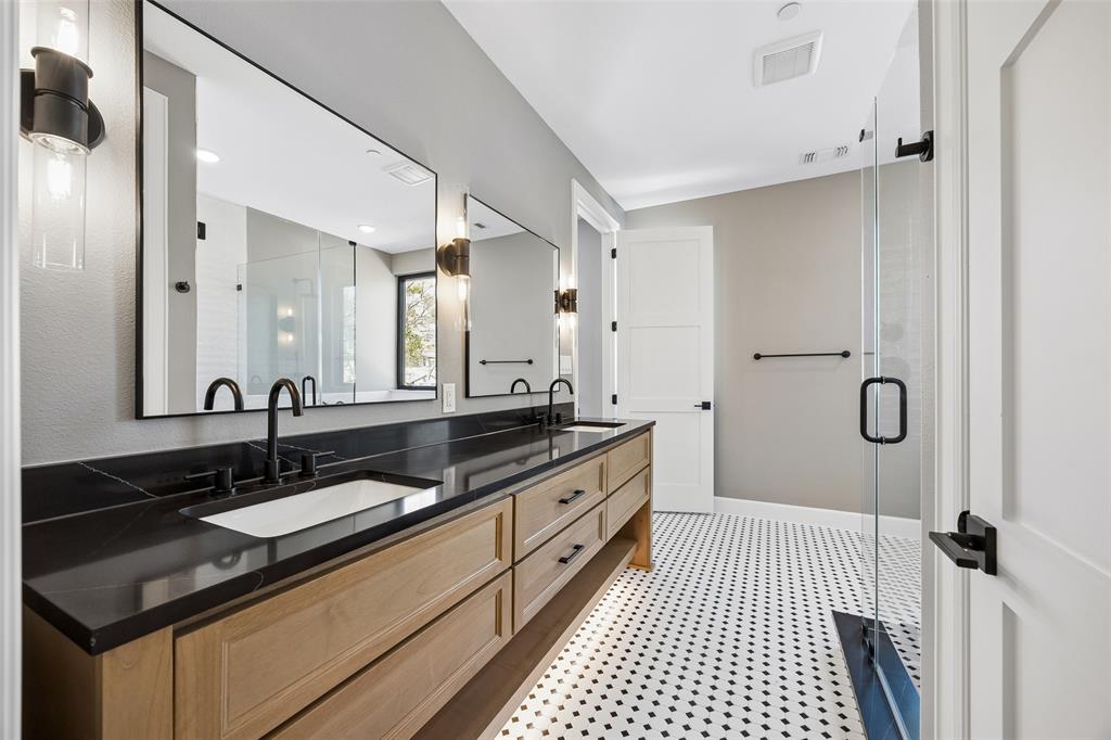 Dual-sink vanity with matte black fixtures, stone-surface countertop, natural wood-finish cabinetry, geometric tile flooring, and a glass-enclosed shower