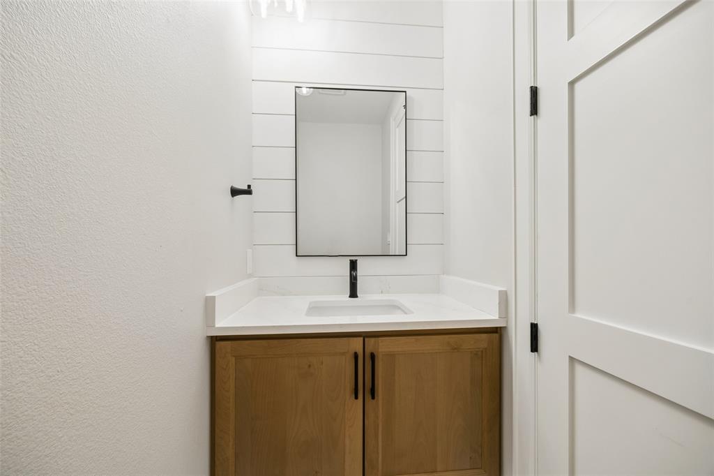 Vanity featuring a light wood-finish cabinet, white countertop with an integrated rectangular sink, shiplap accent wall, framed mirror, and matte black hardware