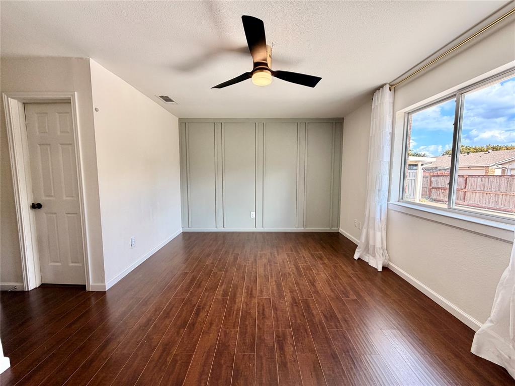 Wood-finish flooring, sage-colored accent wall with vertical molding, three-blade ceiling fan with integrated light fixture, and large pane window