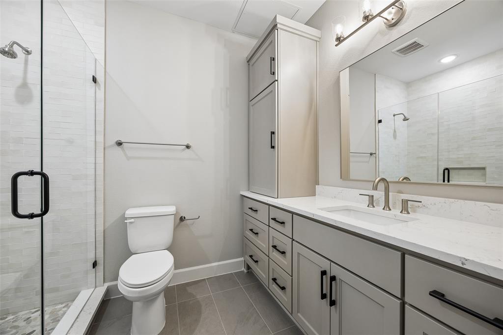 Bathroom featuring a vanity with neutral-toned cabinetry, stone-look countertop, integrated sink, and matte black hardware, accompanied by a glass-enclosed shower with tiled surround, full-width mirror, and built-in vertical storage cabinet