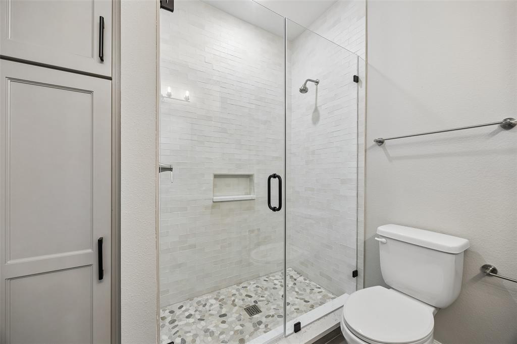 Walk-in shower featuring floor-to-ceiling tile, integrated recessed shelving, a frameless glass door, and a pebble-stone basin floor, complemented by floor-to-ceiling cabinetry with matte black hardware