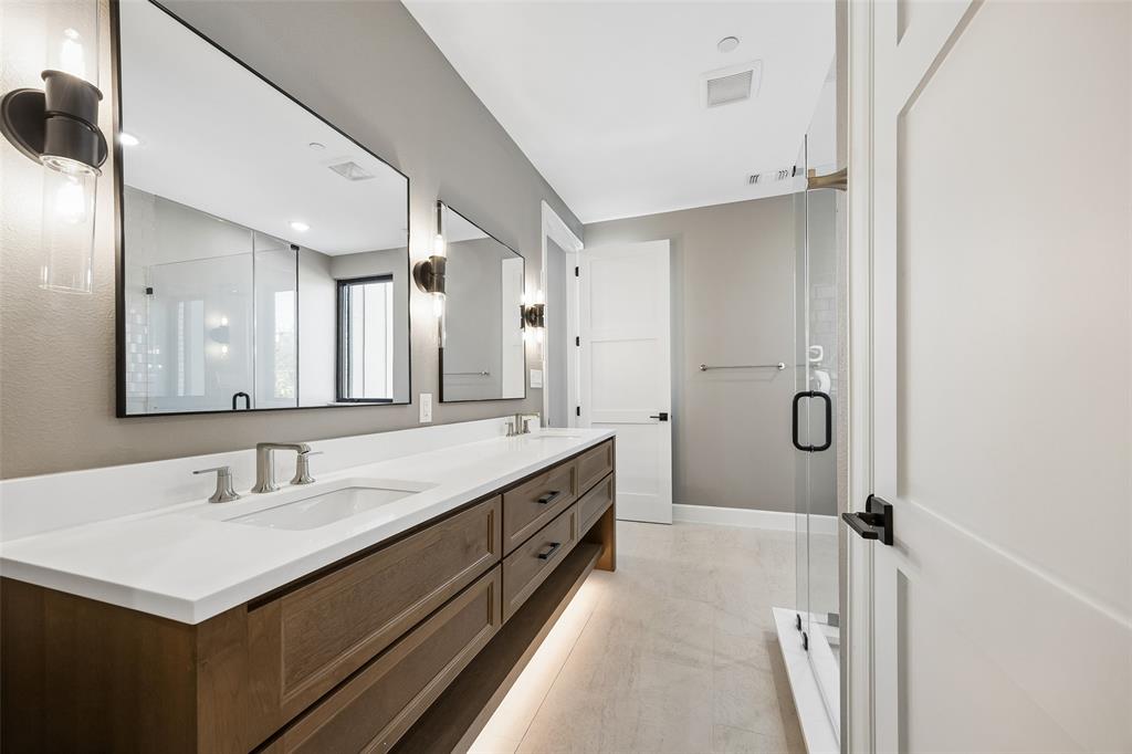 Double vanity with wood-finish cabinetry, white solid-surface countertop, rectangular undermount sinks, under-cabinet lighting, and frameless glass shower enclosure
