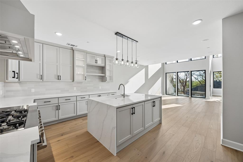 Kitchen featuring light-toned cabinetry, an island with waterfall-edge stone counters, a stainless steel gas range, multi-pendant lighting, and wide-plank wood-finish flooring