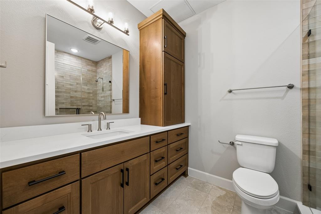 Wood-finish cabinetry featuring an integrated floor-to-ceiling storage unit, expansive white solid-surface countertop, rectangular undermount sink, multi-light vanity fixture, and tiled shower enclosure with glass paneling