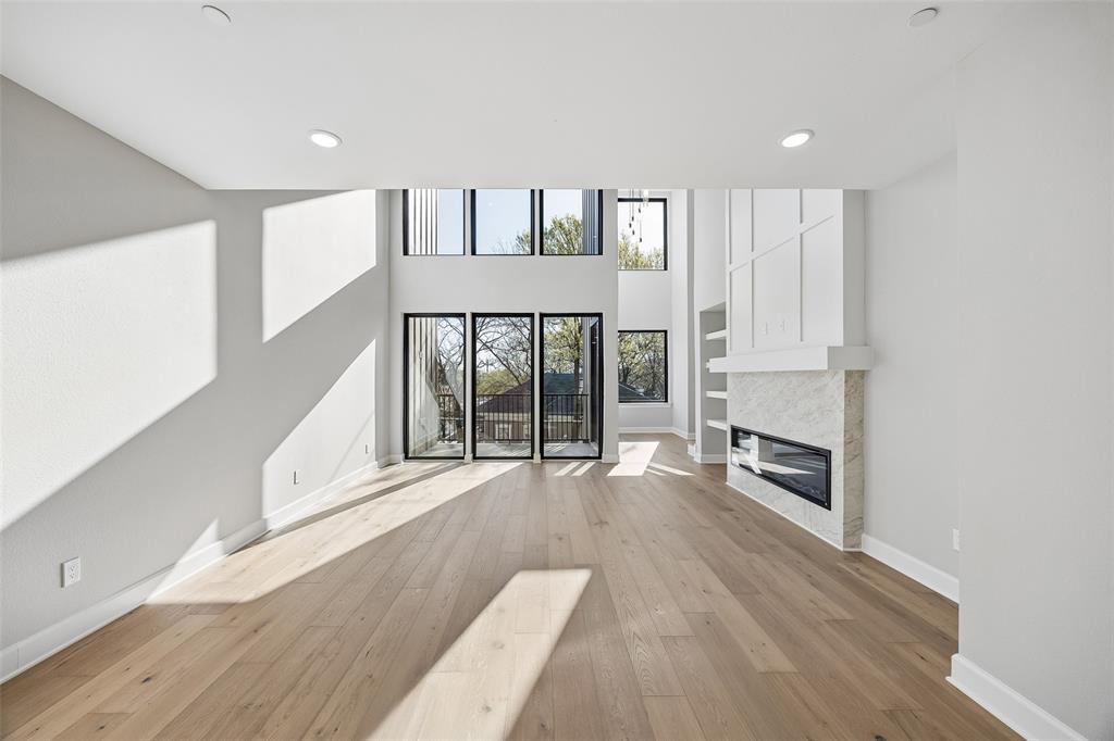 Double-height living area featuring floor-to-ceiling windows, wide-plank wood-finish flooring, recessed lighting, and a linear fireplace with stone-finish surround and built-in vertical millwork