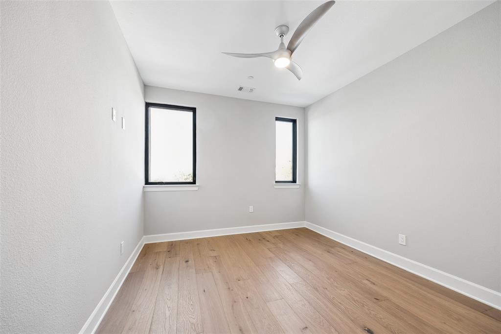 Interior room featuring wood-finish flooring, neutral wall tones, two windows with black frames, and a contemporary ceiling fan with integrated lighting
