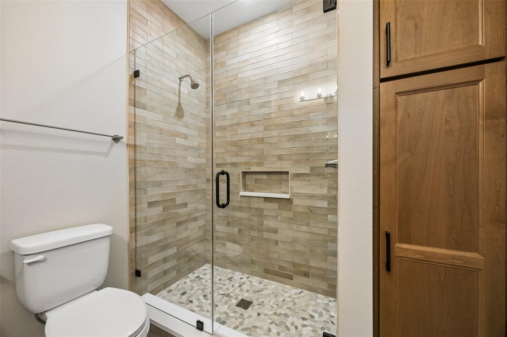 Walk-in shower featuring a frameless glass enclosure, pebble-stone flooring, stacked-stone tile surround, built-in recessed shelving, and a floor-to-ceiling built-in wood-finish cabinet