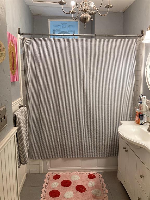Bathroom featuring a white vanity cabinet with a curved integrated sink, beadboard wainscoting, a metal-finish multi-arm chandelier, and a tub enclosure with tiled surround
