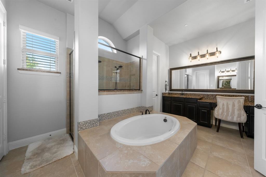 Master Bathroom featuring a tile-surround soaking tub, glass-enclosed shower, dark-finish wood cabinetry, stone-surface dual vanity, and a full-width framed mirror