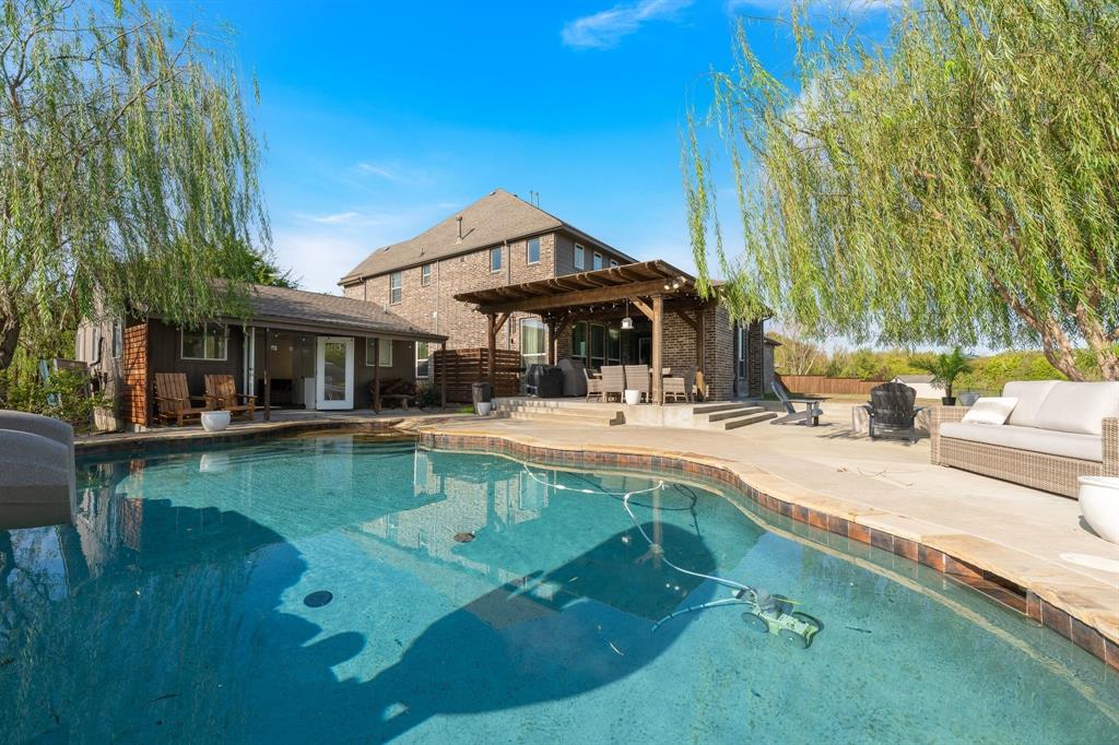 Swimming pool with decorative tile border, expansive concrete patio, timber-framed covered pergola, and mature weeping willow trees