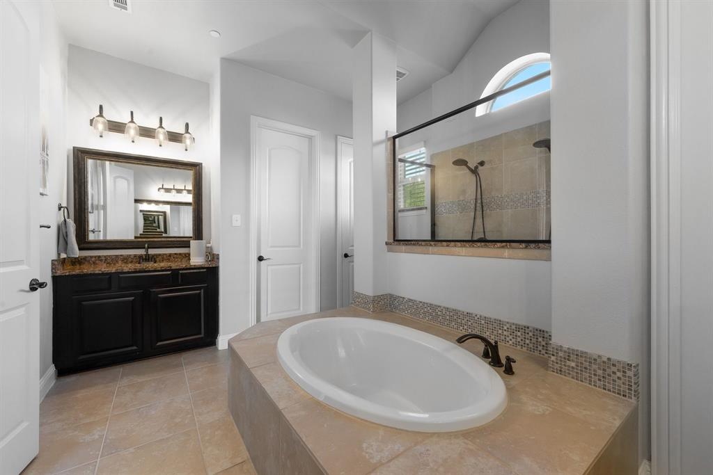 Master built-in soaking tub with tile surround, dark wood vanity, stone countertop, arched window, and glass-enclosed shower