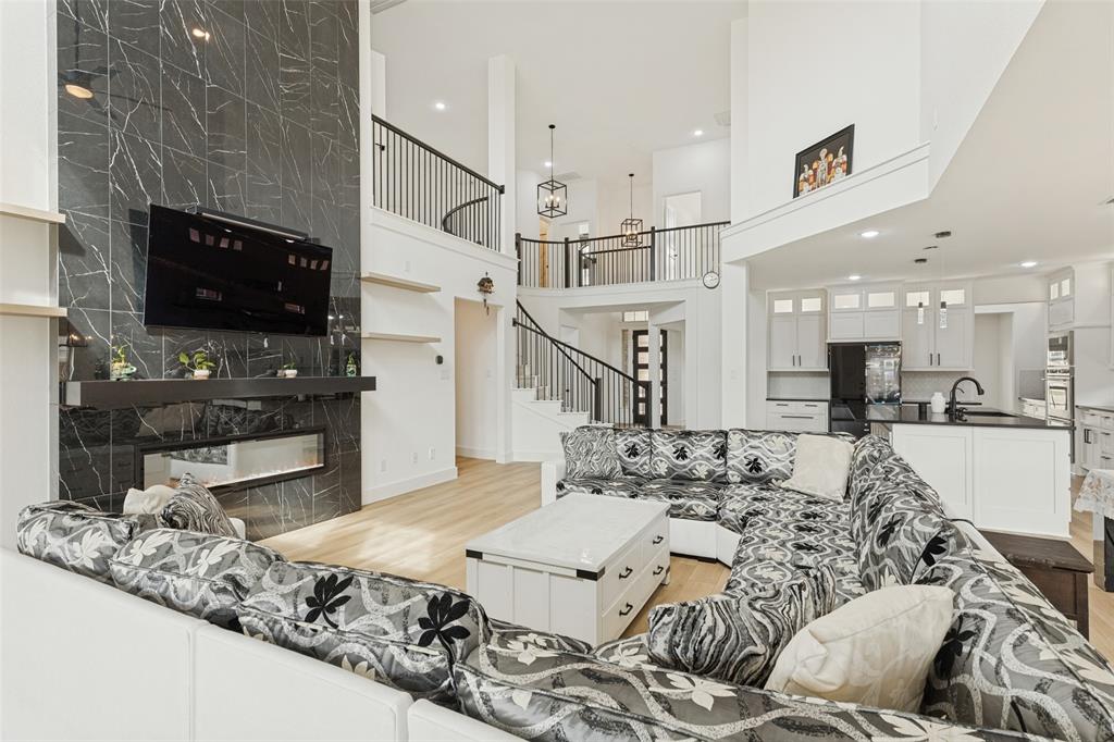 Two-story living area featuring a full-height dark stone-tiled fireplace, wood-finish flooring, white cabinetry with integrated appliances, and a wrought-iron staircase railing