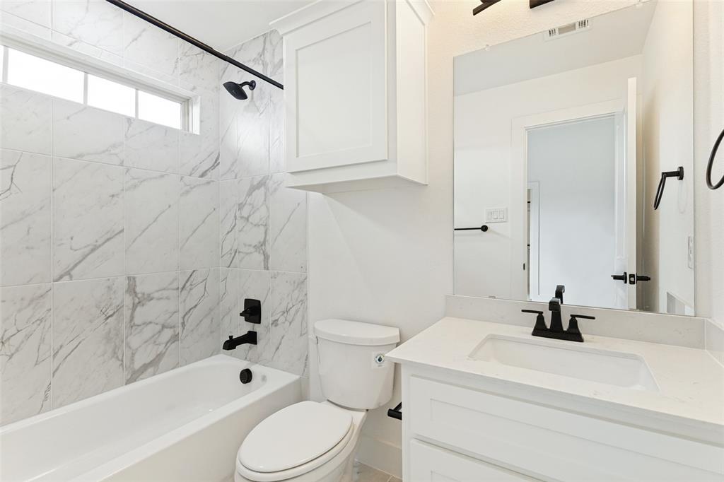 Bathroom featuring a shower-tub combination with stone-patterned wall tile, a white vanity with a light-toned countertop, a rectangular undermount sink, matte black hardware, and an overhead cabinet
