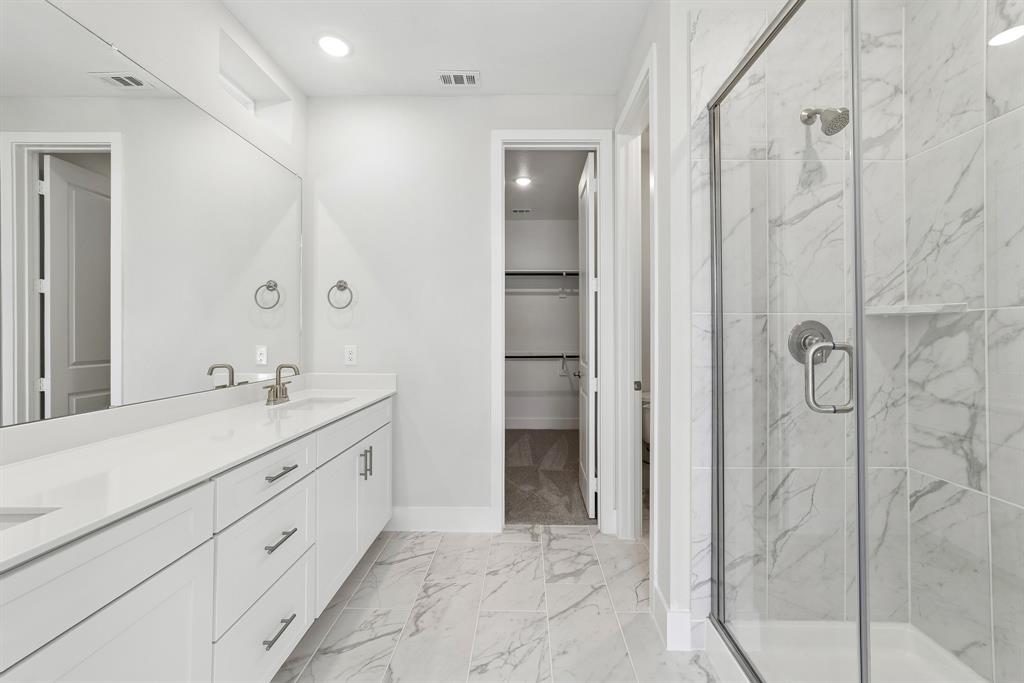 Bathroom featuring a dual-sink vanity with white cabinetry and solid-surface countertops, walk-in shower with floor-to-ceiling tile surround, and tile flooring throughout