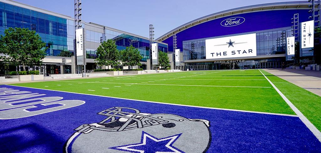 Synthetic turf athletic field featuring professional markings, paved pedestrian walkways, expansive glass-facade exterior structures, and integrated stadium lighting Synthetic turf athletic field featuring professional markings, paved pedestrian walkways, expansive glass-facade exterior structures, and integrated stadium lighting