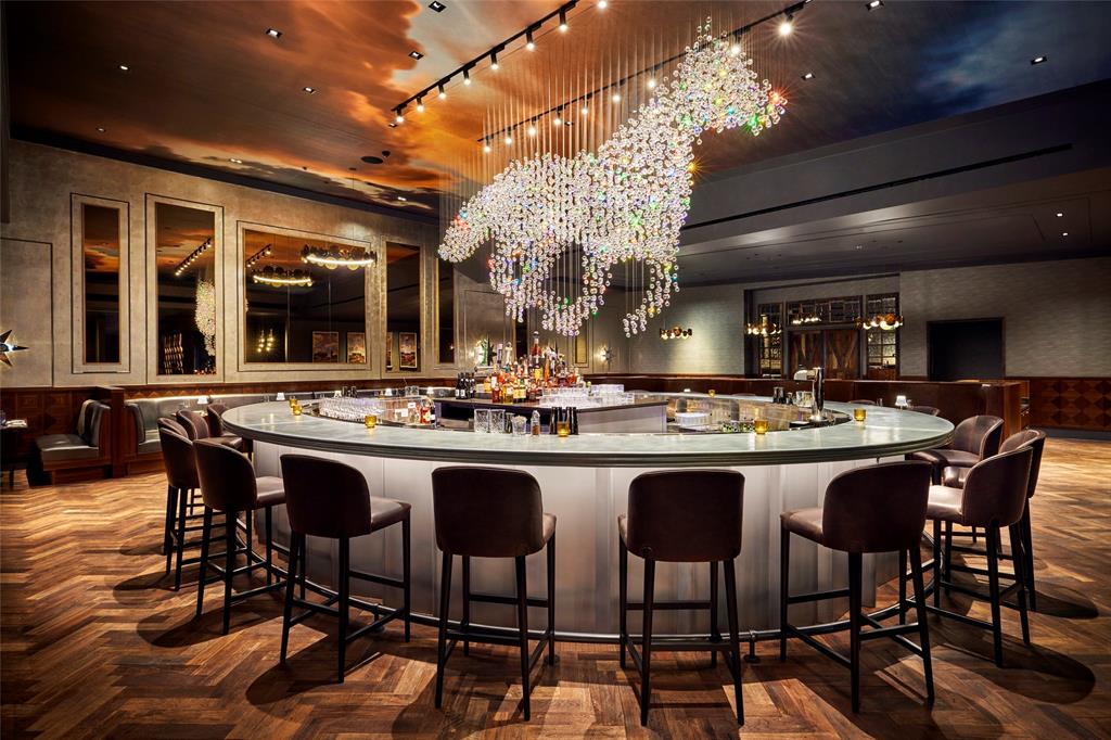 Trick Rider at FRISCO PGA Circular bar featuring a translucent base, herringbone-patterned wood flooring, custom crystal light fixture, wall-mounted recessed mirrors with decorative trim, and an expansive cloud-patterned ceiling mural