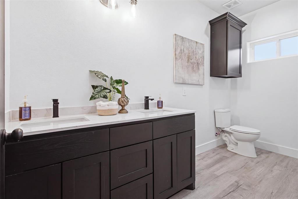 Double vanity featuring dark-toned cabinetry, light-colored stone countertops with integrated rectangular sinks, matte black faucets, and wood-finish flooring, complemented by an overhead storage cabinet