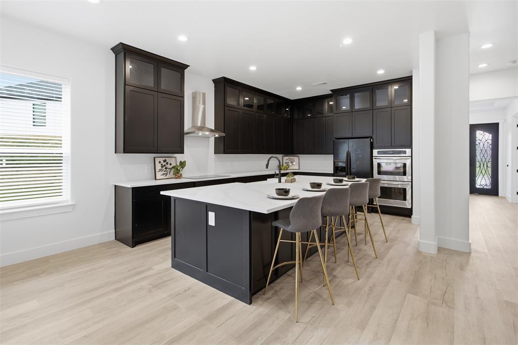 Kitchen featuring dark cabinetry with glass-fronted upper units, a large central island with a stone-surface countertop, integrated stainless steel double wall ovens, a stainless steel range hood, and light-toned wood-finish flooring