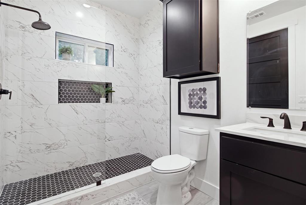 Walk-in shower with floor-to-ceiling stone-pattern tile, contrasting dark hexagonal floor tile, built-in wall niche, matte black rain-style shower head, and vanity with dark cabinetry