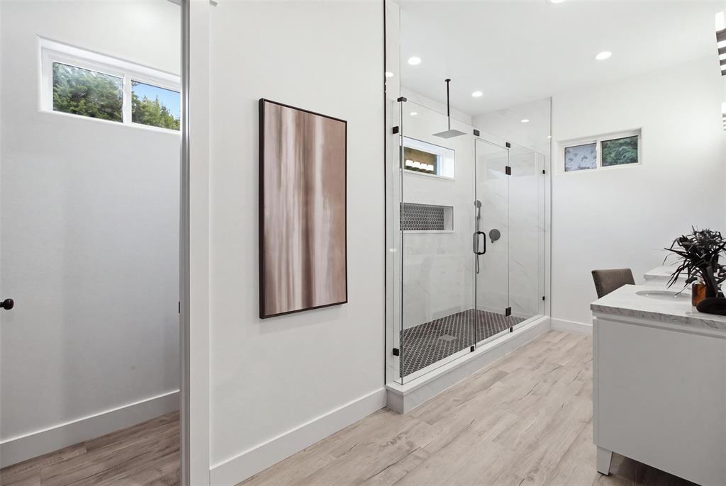 Walk-in shower with a glass enclosure, rainfall showerhead, and mosaic tile flooring, complemented by a vanity with a stone-finish countertop and wood-finish flooring
