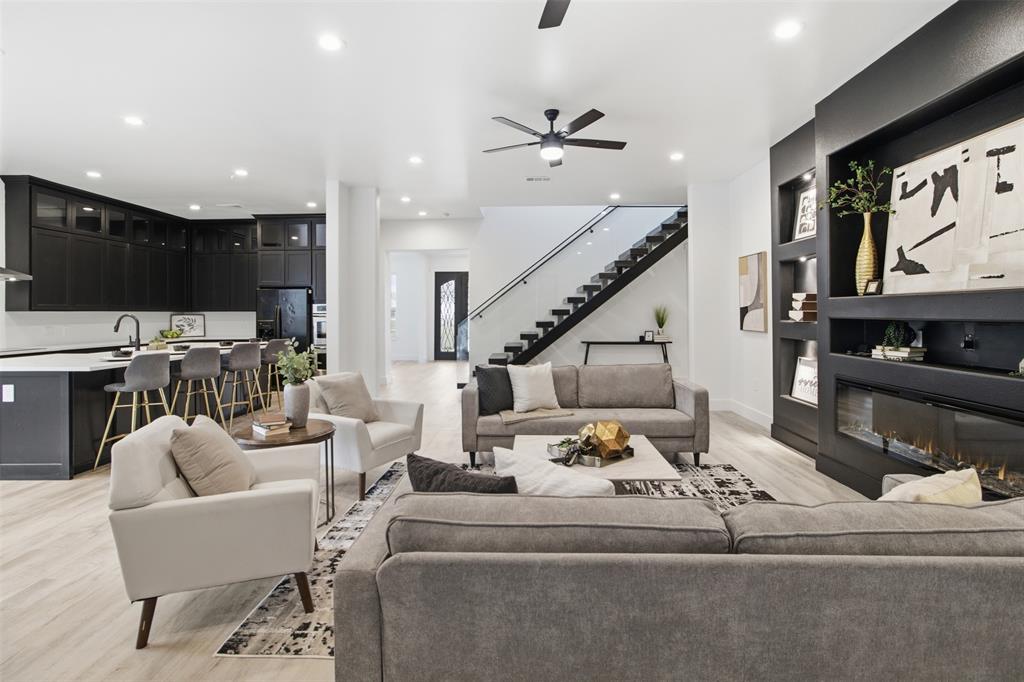 Open-concept living space featuring wood-finish flooring, a kitchen with dark cabinetry and a central island, a built-in wall unit with an integrated linear fireplace, and a staircase with a glass-panel railing