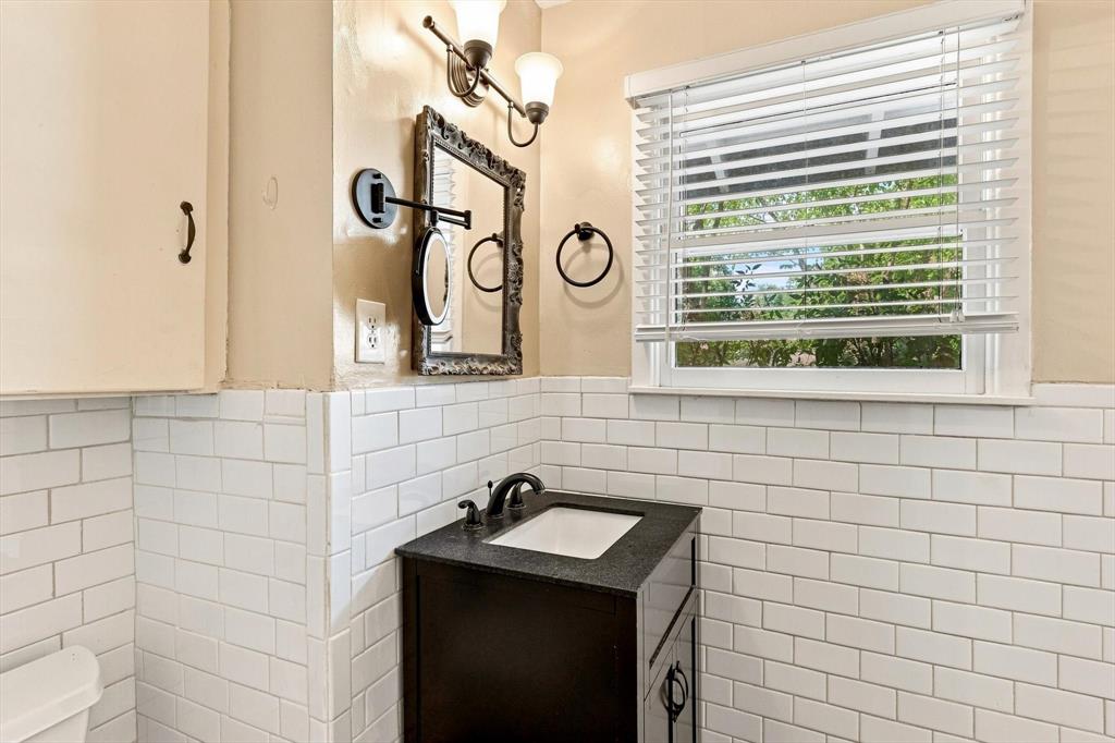 White subway tile wainscoting, dark finish vanity with a stone countertop, integrated undermount sink, multi-light vanity fixture, and horizontal blind window treatment