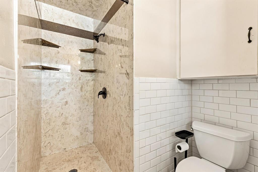 Shower enclosure featuring stone-finish wall surrounds, built-in shelving, matte black fixtures, and white subway tile wainscoting