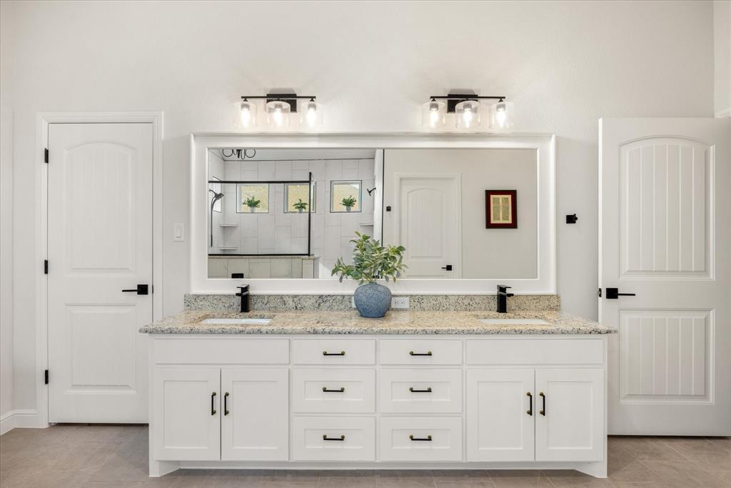 Dual-sink vanity featuring stone countertop, white cabinetry with matte black hardware, large framed mirror, and dual wall-mounted light fixtures
