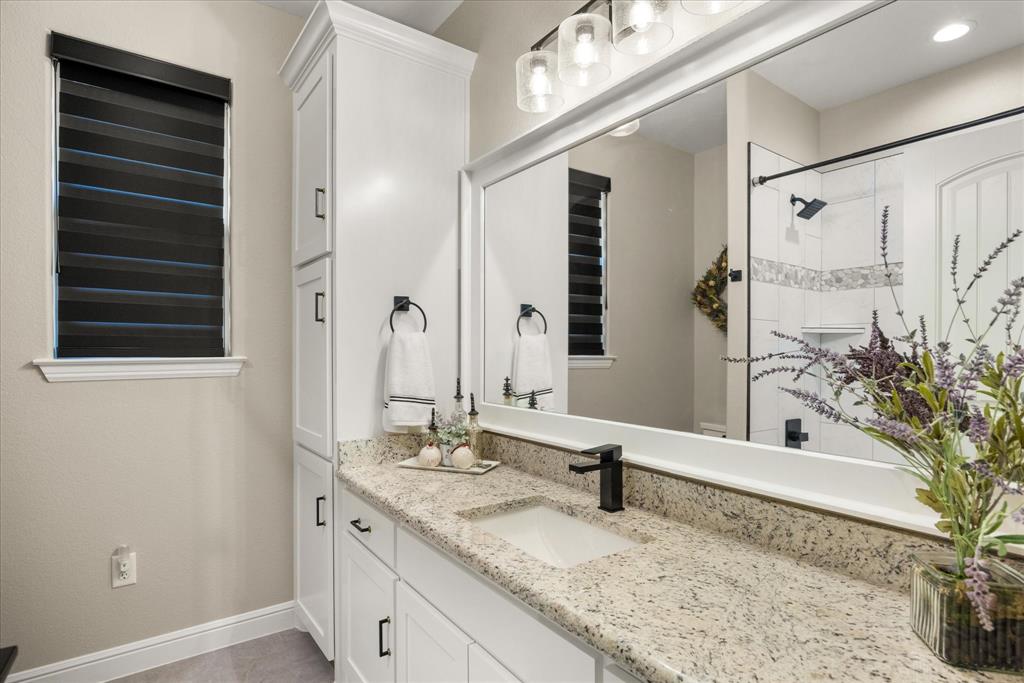 Bathroom featuring white cabinetry with a floor-to-ceiling storage tower, stone countertop, matte black faucet, full-width framed mirror, and walk-in shower with tiled surround